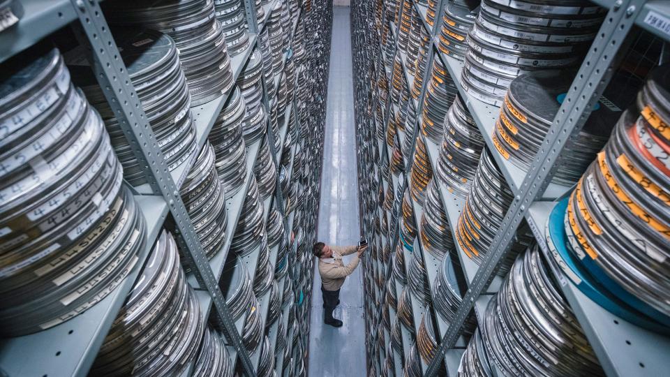 A photo from inside the BFI National Archive. An archive worker is shown standing between two tall walls of shelves containing film reel cannisters.